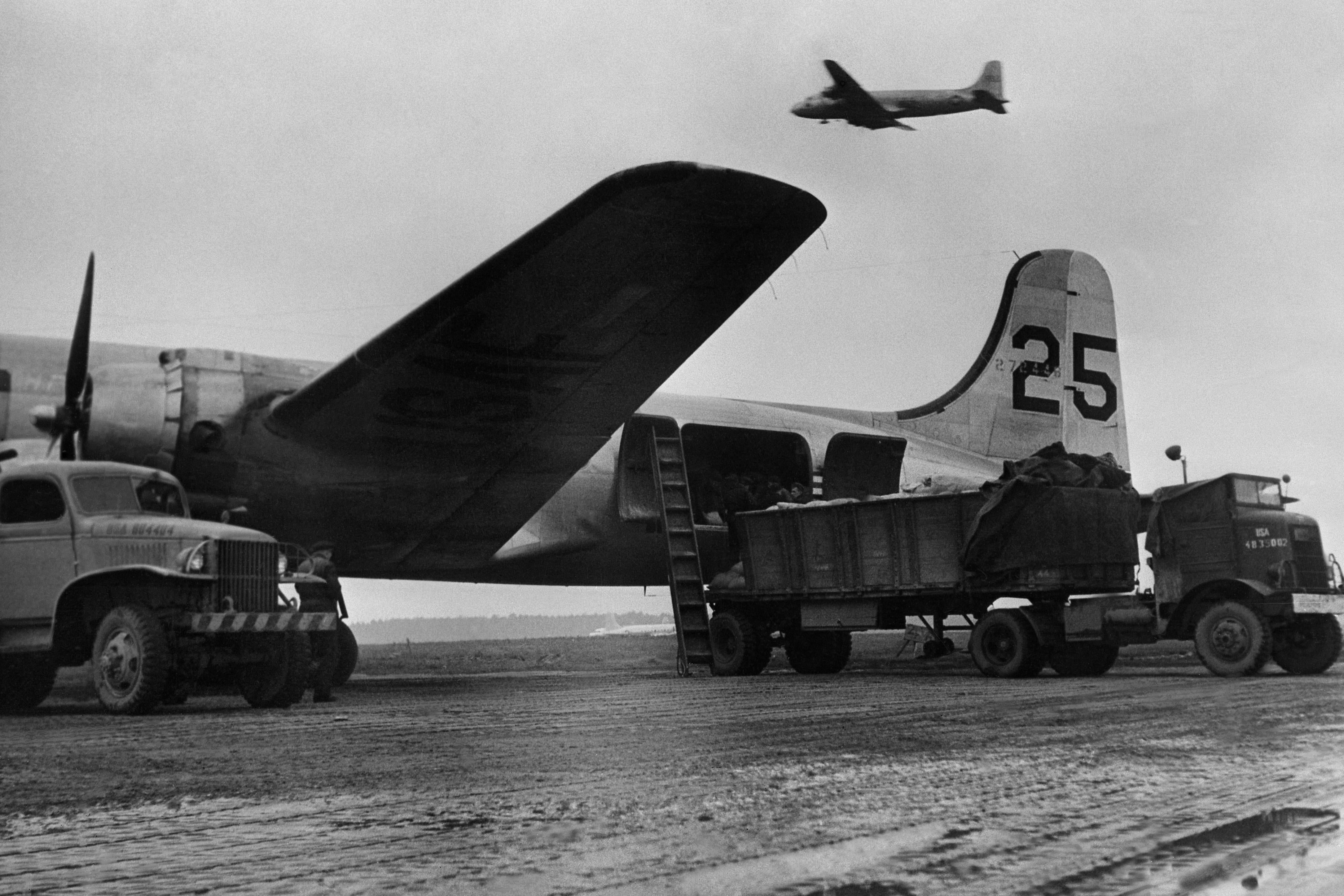Trucks load planes on a tarmac as another plane flies by.