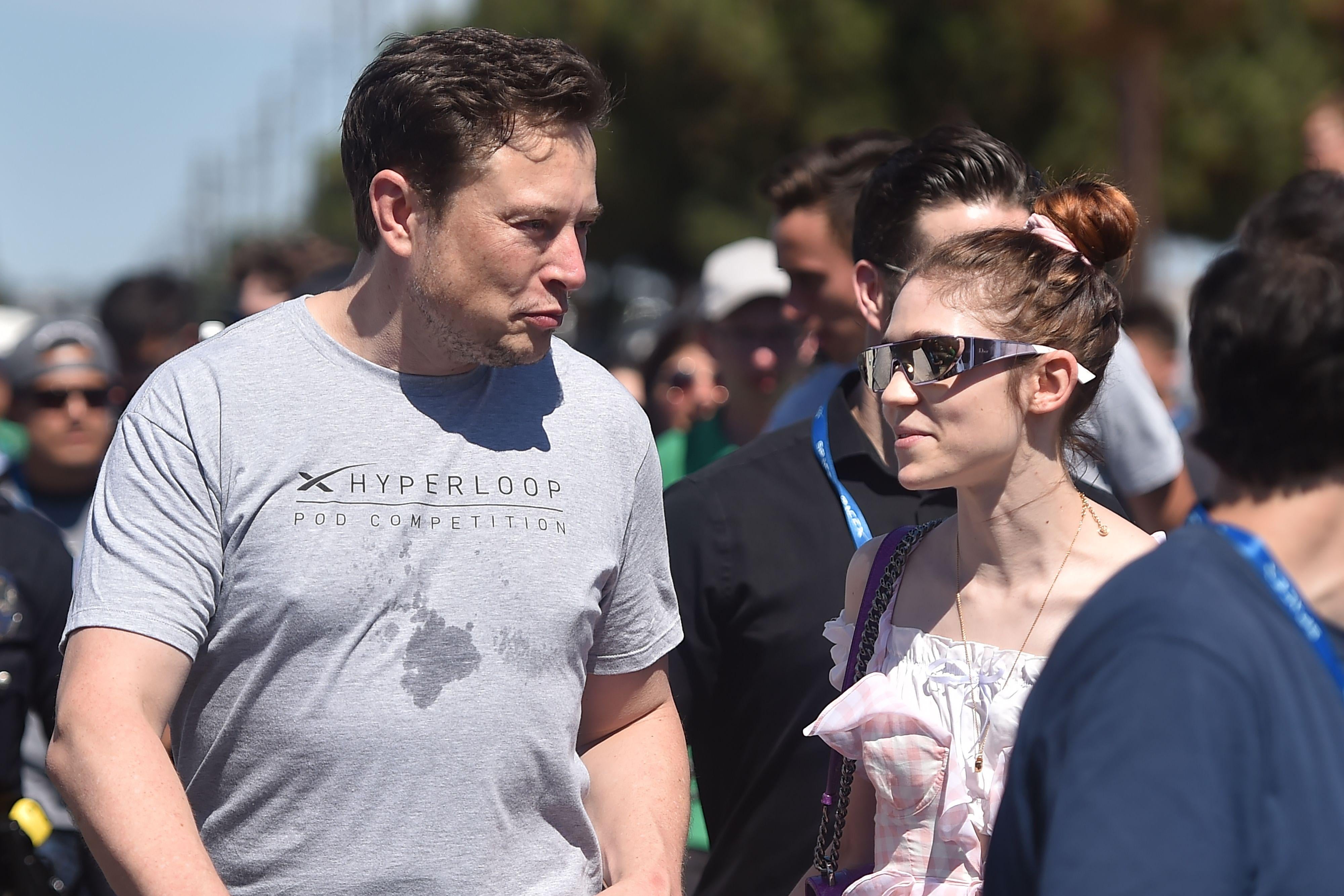Musk in a Hyperloop T-shirt and Grimes wearing futuristic metal sunglasses and a dress walk next to each other in a crowd outside
