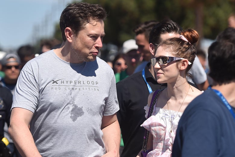 Musk in a Hyperloop T-shirt and Grimes wearing futuristic metal sunglasses and a dress walk next to each other in a crowd outside