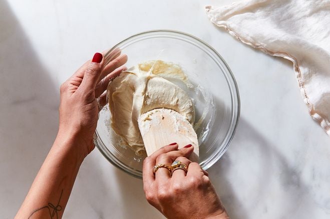 Hands use a wooden spatula to knead dough in a glass bowl.